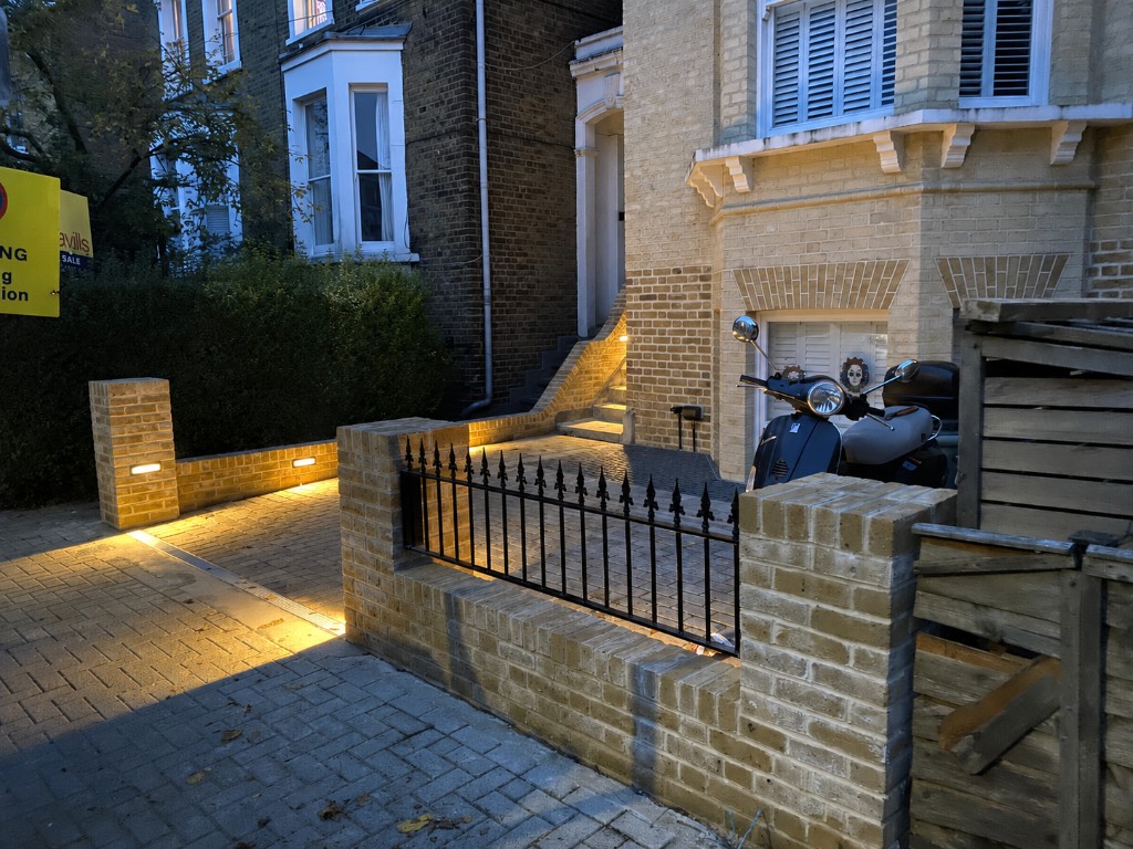 Buff brick walls and pillars with integrated warm recessed lighting beside block paving at dusk