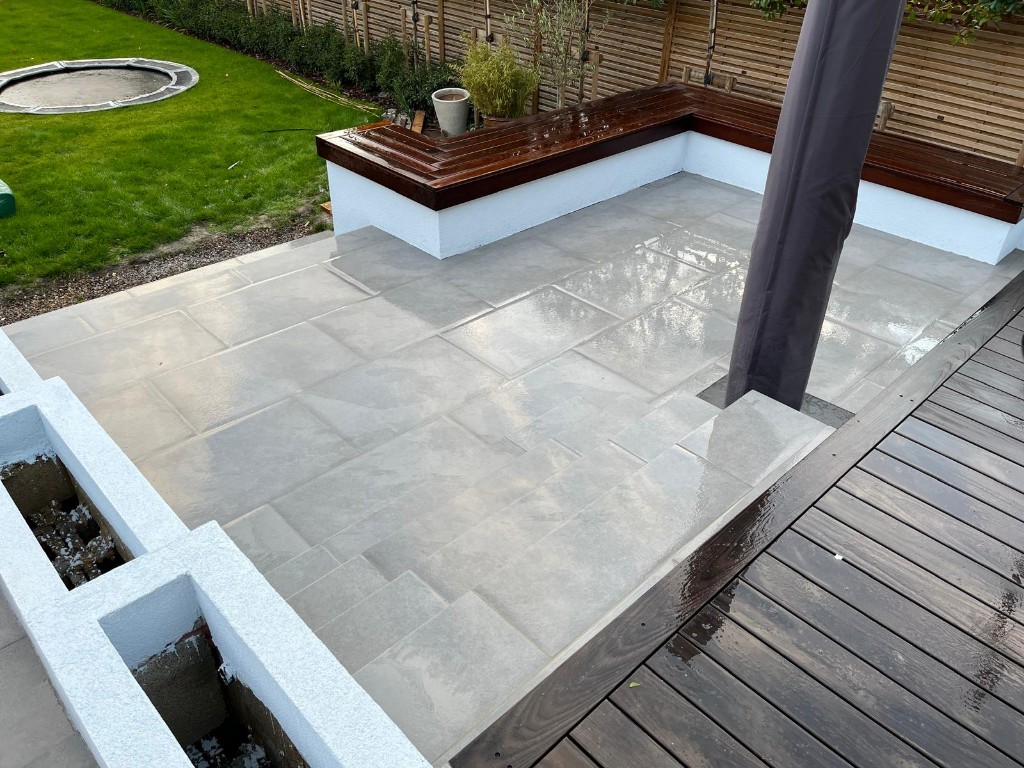 Large grey stone patio with built-in seating, decking and lawn beyond horizontal fencing
